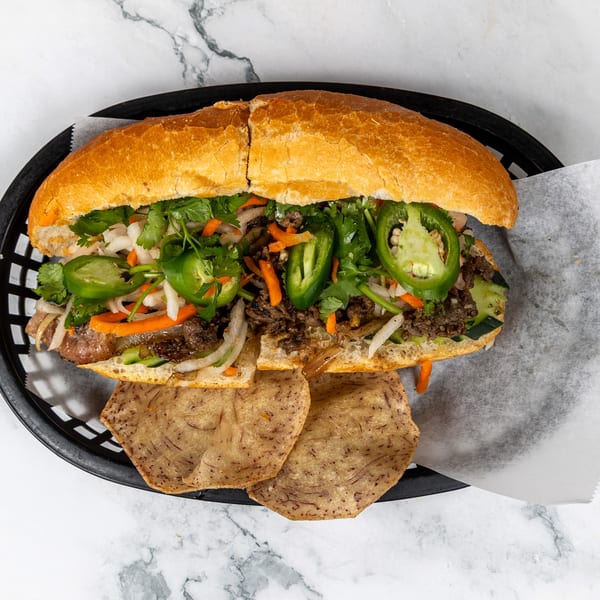 A Vietnamese Bánh Mì sandwich with meat, cilantro, jalapeños, and pickled vegetables, served with chips.