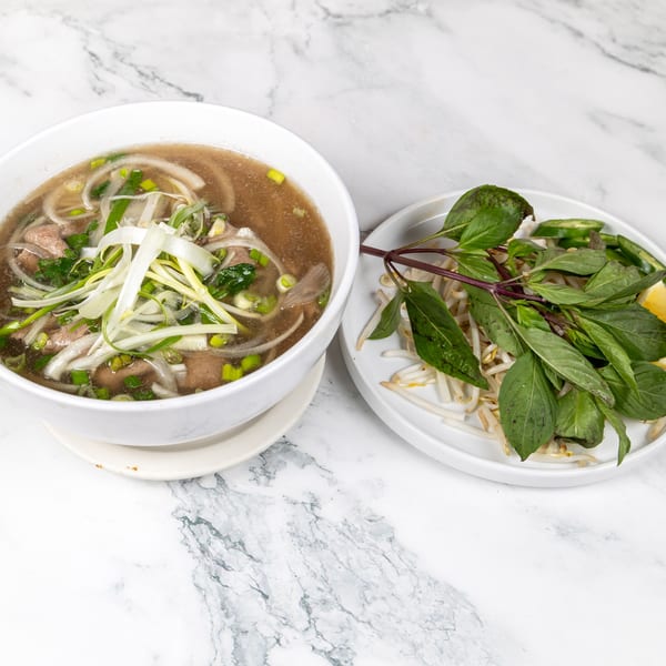 A bowl of Vietnamese pho with beef, onions, and scallions, served with a side of fresh herbs.